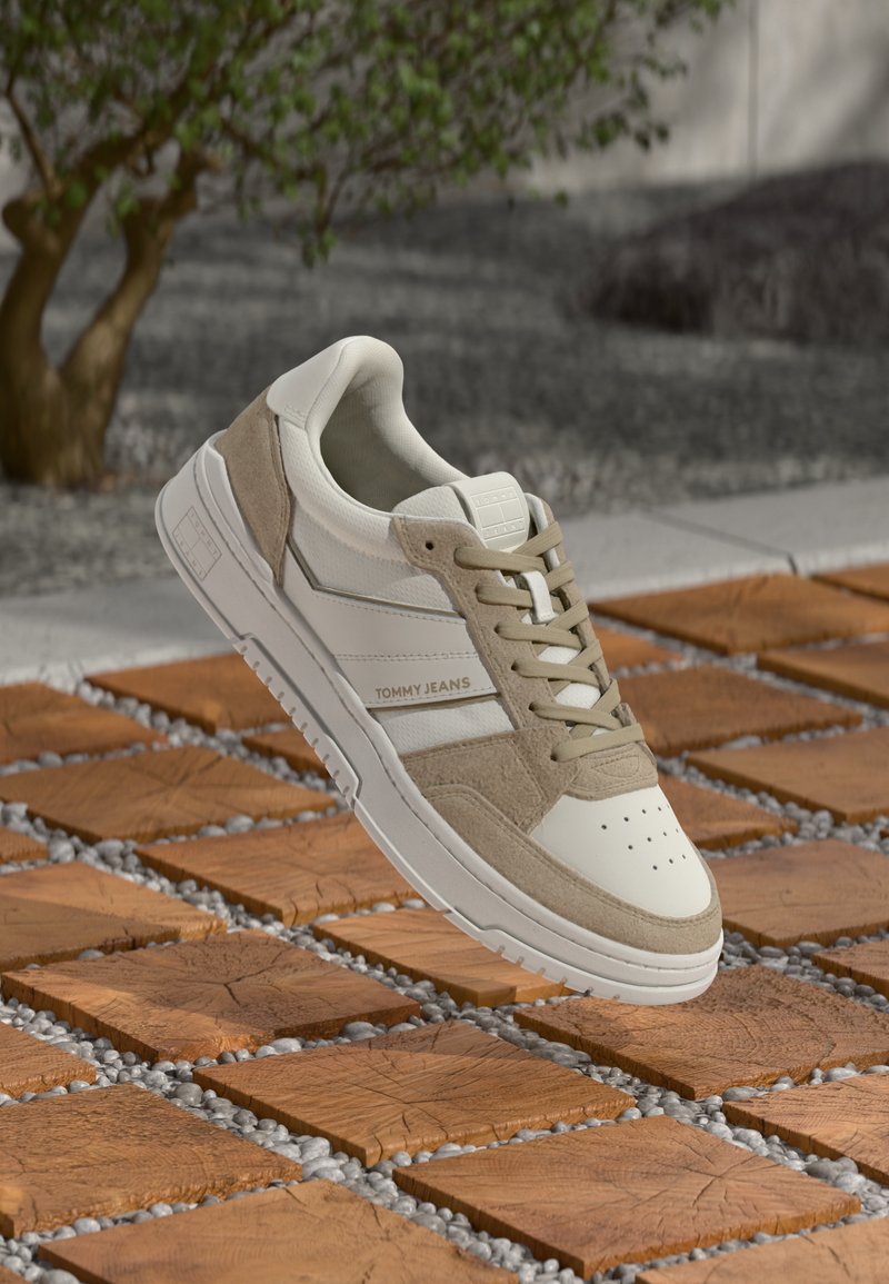White and beige Tommy Jeans sneaker with laces, floating above a wooden tile and pebble outdoor floor with a tree in the background.