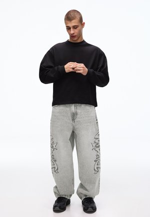 Junger Mann schaut nach unten, trägt ein schwarzes Langarmshirt, weite graue Jeans mit Stern- und Graffiti-Designs sowie schwarze Turnschuhe, steht vor weißem Hintergrund.