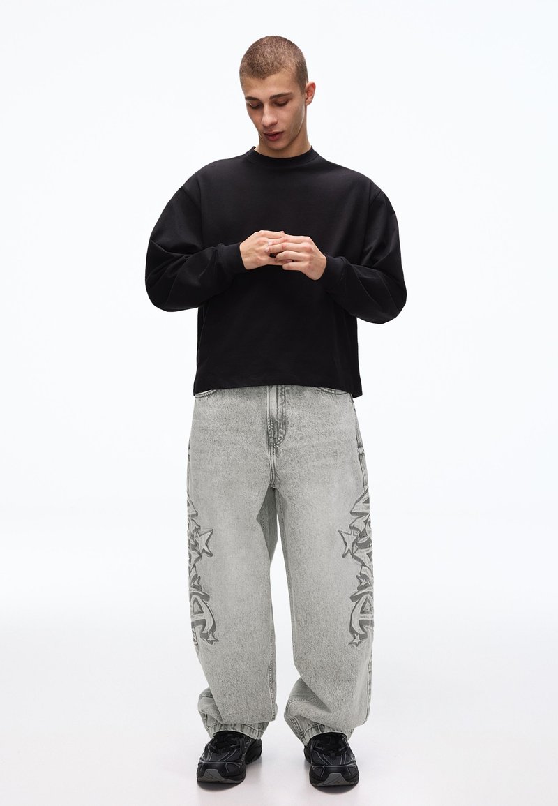 Jeune homme regardant vers le bas, portant un t-shirt noir à manches longues, un jean gris ample avec des motifs d'étoiles et de graffiti, et des baskets noires, debout sur un fond blanc.