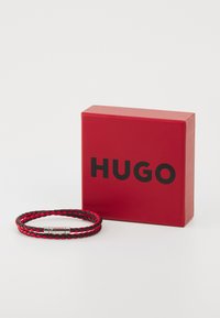 Schwarzes und rotes geflochtenes Lederarmband mit Metallverschluss, das neben einer roten Box mit dem schwarzen Logo "HUGO" präsentiert wird.
