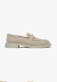 Beige suede loafers with a decorative woven strap, round toe, and a low, chunky cream sole. Stitching accents along the edges.