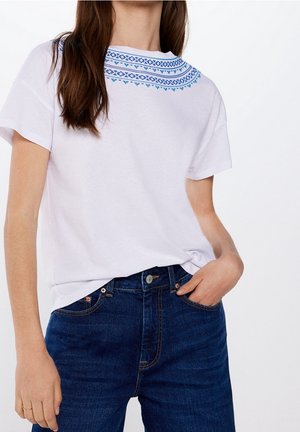 Hvid bomulds-T-shirt med blåt broderet mønster rundt om den runde halsudskæring, kombineret med mørkeblå denim jeans. Enkelt ærmedesign.