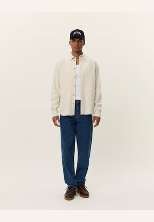 Chemise crème à boutons sur un débardeur blanc, associée à un pantalon en denim bleu et des chaussures marron. Casquette noire avec le logo "LES DEUX".
