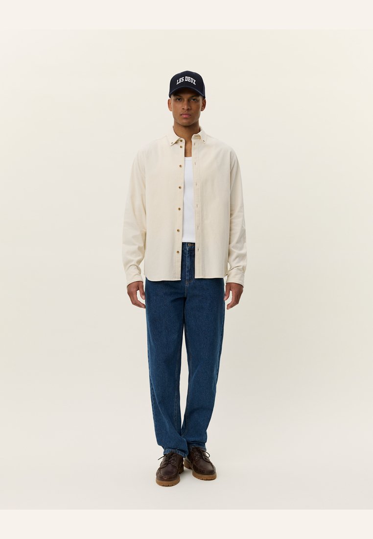 Cream button-up shirt over a white tank, paired with blue denim pants and brown shoes. Black cap displays "LES DEUX" logo.