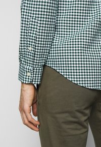 Zelená a bílá kostkovaná košile s manžetami na knoflíky, doplněná o khaki kalhoty. Látka působí texturovaně a lehce.