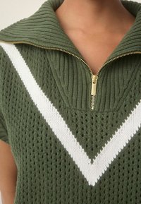 Pletený svetr v olivově zelené barvě s vysokým límcem a zlatým zipem. Obsahuje bílo-červený V-čtvercový vzor přes hruď a strukturované vrstvení.