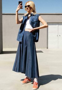 Chemise sans manches en denim sur un top blanc cropped, associée à une longue jupe en denim et des baskets orange avec des rayures blanches. Fond gris.