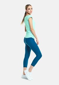 Lichtteal kleurig, aansluitend T-shirt met korte mouwen, gecombineerd met donkerteal kleurige capri-legging en witte sport sneakers. Gladde texturen, nauwsluitend ontwerp.