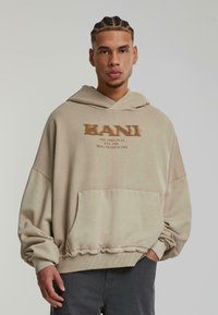 Sweat-shirt à capuche beige avec une coupe décontractée, doté d'une poche avant, de poignets côtelés et d'un logo imprimé en texte marron foncé.