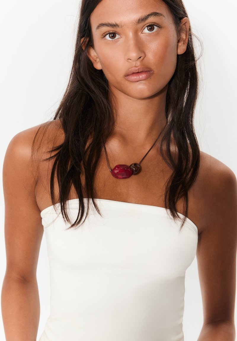 Naszyjnik z czerwonych i brązowych koralików na ciemnym sznurku, z nieregularnymi kształtami i wypolerowaną fakturą, noszony na białym, bezramkowym topie.