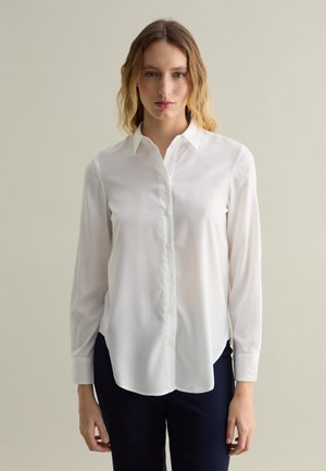 Weißes Hemd mit langen Ärmeln, aus glänzendem Baumwollstoff, mit einem geraden Kragen und abgerundetem Saum, kombiniert mit marineblauen Hosen.