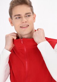 Rote Fleece-Weste mit Reißverschluss und hohem Kragen, glatter Textur und einem kleinen Reißverschlussdetail. Getragen über einem weißen Langarmshirt.