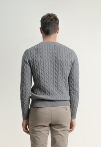 Suéter gris de punto trenzado con dobladillo y puños acanalados, que presenta un patrón texturizado. Combinado con pantalones de color claro, mostrando un ajuste relajado.