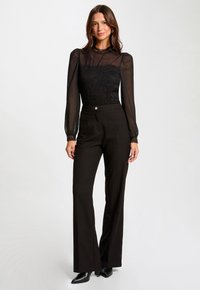 Body en dentelle noire avec des manches longues transparentes, associé à un pantalon noir taille haute. Le pantalon présente un bouton à l'avant et un design élégant.