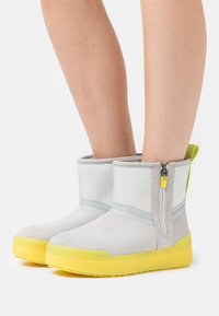 Ljusgrå ankelboots med gula sulor och sidodragkedjor, bärs på bara ben, visade mot en vit bakgrund.