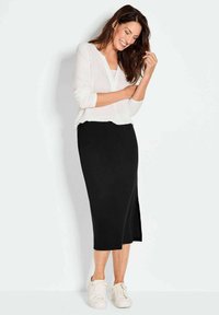 Schwarze knielange Rock mit einem seitlichen Schlitz, kombiniert mit einer weißen Bluse mit langen Ärmeln. Model trägt weiße Sneakers und steht vor einem hellen Hintergrund.