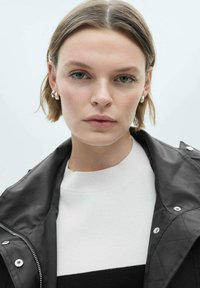 Black quilted jacket with silver hardware, layered over a white and black striped knit sweater. Short hair, minimal makeup, neutral background.