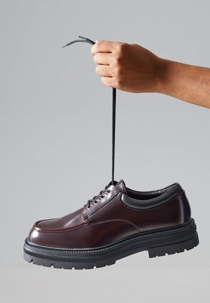 Chaussure en cuir bourgogne avec un bout arrondi, semelle épaisse en caoutchouc noir et lacets gris, tenue par le dessus.