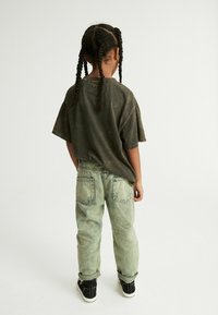 Un niño lleva una camiseta de manga corta, holgada y de color verde oscuro, junto con unos vaqueros ajustados y de color azul claro con una textura deslavada, complementados con zapatillas negras.