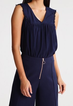 Vrouw draagt een mouwloze marineblauwe blouse met gerimpelde randen en marineblauwe hooggetailleerde broek met een voorste ritssluiting met ringtrekker.