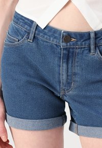 Tamne traperice tamnoplave boje s prevrnutim rubom, s prednjim džepovima, srebrnim zatvaračem i detaljima u kontrastnoj boji.