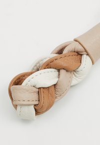Accessoire en cuir tressé dans des tons beige, marron et blanc avec des coutures visibles. Texture douce et finition lisse avec une forme arrondie.