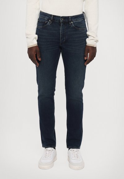 Jeans en denim bleu foncé avec une coupe ajustée, un design à cinq poches et un léger effet de délavage. Associés à des baskets blanches à lacets.