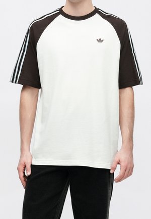 Mann trägt ein weißes und schwarzes Adidas T-Shirt mit drei weißen Streifen auf schwarzen Ärmeln und schwarzen Hosen, steht vor einem einfarbigen Hintergrund.