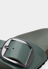 Πράσινο σανδάλι slide με γυαλιστερό φινίρισμα, που διαθέτει μια μεγάλη ασημένια αγκράφα. Η σόλα είναι καφέ και υφασμένη, τονίζοντας τον χαλαρό του σχεδιασμό.