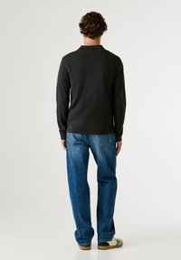 Černá košile s dlouhým rukávem a jednoduchým límečkem, v kombinaci s volnými modrými denimovými džíny a béžovými teniskami, pohled zezadu.