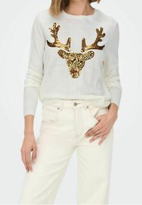 Witte gebreide trui met een gouden pailletten rendierontwerp op de voorkant, gecombineerd met lichtgekleurde jeans. De textuur is soepel en zacht.