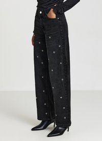 Black high-waisted wide-leg jeans with silver eyelet details throughout; fabric is denim with a soft texture. Paired with pointed black heels.