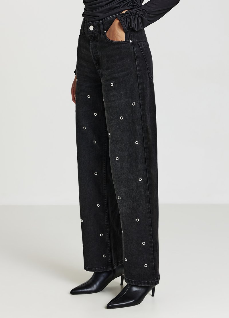 Black high-waisted wide-leg jeans with silver eyelet details throughout; fabric is denim with a soft texture. Paired with pointed black heels.