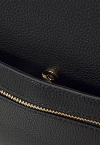 Černá texturovaná kožená taška uvnitř se zlatým zipem a kulatým magnetickým uzávěrem, ukazující detaily šití a struktury.