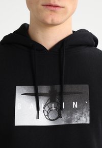 Primo piano di una persona che indossa un felpa nera con una grafica di un canestro da basket in bianco e nero e la parola "BALLIN'" sopra il design.