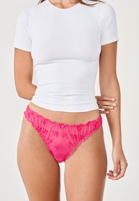 Pink sheer thong with ruffled edges and a button detail, worn under a fitted white t-shirt. Soft texture and lightweight material.