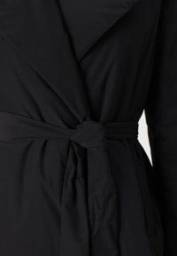Black wrap coat made of smooth fabric, featuring a large collar and a tied waist detail, creating a structured shape with soft draping.