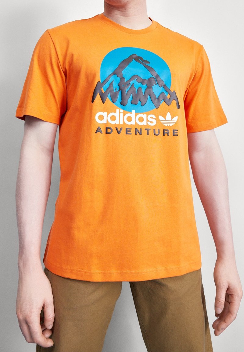 Oranžové bavlněné tričko s modrým kruhovým motivem hor a černým nápisem "adidas ADVENTURE". Krátké rukávy a kulatý výstřih.