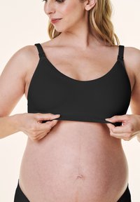 Czarny stanik ciążowy z regulowanymi ramiączkami i miękkiego materiału, zaprojektowany dla wygody. Modelka delikatnie unosi pasek stanika.