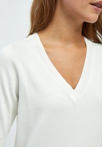 Suéter blanco de cuello en V con textura de punto, mangas largas y bordes acanalados, que muestra un diseño limpio y simple. Sin patrones ni acentos visibles.