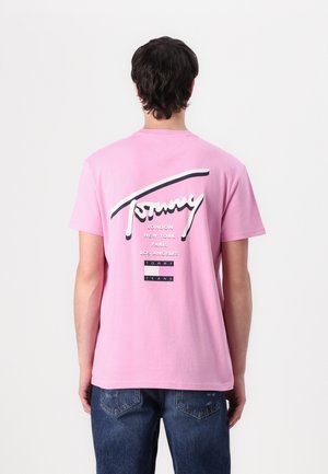 Ružičasta majica s kratkim rukavima s "Tommy" logom u bijelom kurzivnom pismu na leđima, s imenima gradova i detaljima branda u crnim i bijelim akcentima.