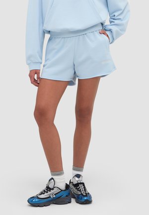 Person iført lyseblå shorts og sweatshirt, hvide sokker og blå og sorte New Balance-sneakers, står med den ene hånd i lommen.