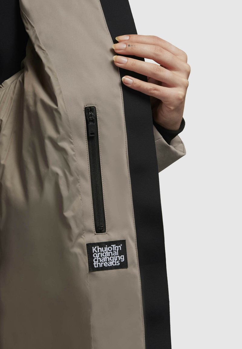 Leichte Jacke aus beige und schwarzem Stoff; verfügt über eine Reißverschlusstasche, hat eine glatte Textur und ein Etikett mit der Aufschrift "Khujo Original Changing Threads."