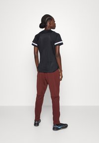 Zwart sportshirt met witte strepen op de mouwen, gecombineerd met losse bordeauxrode broeken en zwarte sportschoenen met blauwe accenten.