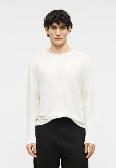 Young man with dark curly hair wearing a long-sleeve white sweater and black pants, standing against a plain light background.