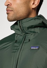 Jachetă verde impermeabilă cu guler înalt, având un fermoar și un patch cu logo-ul Patagonia pe partea stângă. Textură netedă, material ușor.