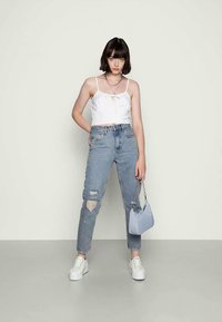Top branco cropped com detalhe de laço, jeans rasgado azul claro, sapatilhas brancas e uma pequena bolsa azul claro. Fundo simples.