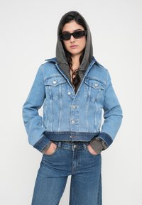 Jachetă din denim albastru deschis, cu design scurt, două buzunare frontale și glugă gri. Purtată cu blugi asortați, albastru.