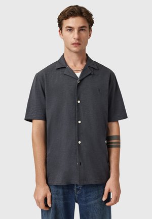 Chemise à manches courtes, gris foncé, à boutons avec un col camp, texture légère et boutons clairs contrastants, portée par-dessus un T-shirt blanc.