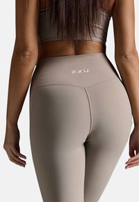Beige Sportleggings mit einem gerippten Bund und Logodetail auf der Rückseite. Das Material scheint dehnbar und figurbetont zu sein.